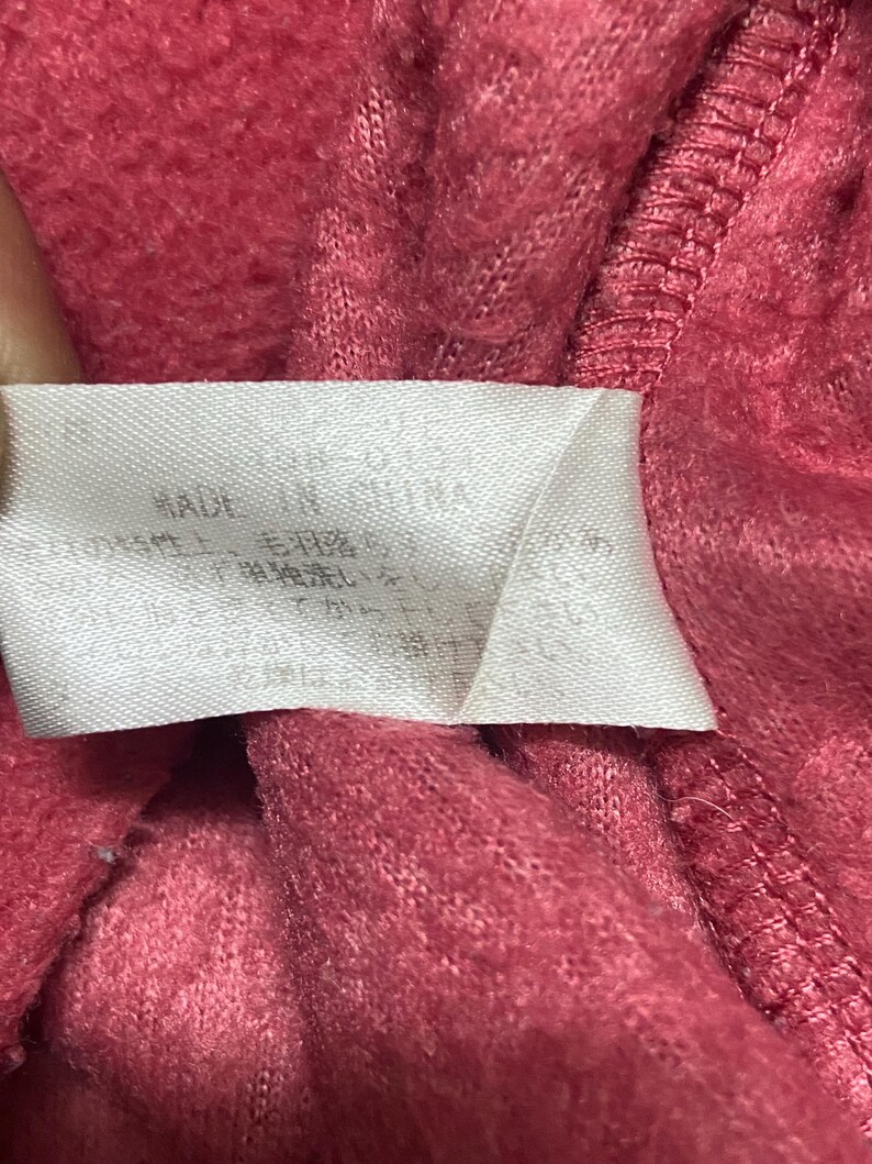 K&ouml;nnte beinhalten: Nahaufnahme eines rosa Stoff-Etiketts mit wei&szlig;em Text, der "MADE IN CHINA" lautet, und anderem Text in einer Fremdsprache.