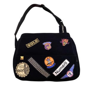 Könnte beinhalten: Schwarze Umhängetasche mit mehreren Aufnähern. Aufnäher enthalten das Wort "GOLD", einen Tiger und das Wort "Numéro". Die Tasche hat einen langen schwarzen Riemen und einen kleinen goldenen Nieten.