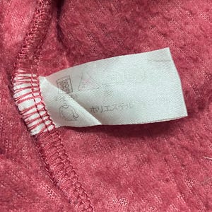 K&ouml;nnte beinhalten: Ein wei&szlig;es Etikett mit schwarzem Text und einer kleinen Grafik einer Person, die sich die H&auml;nde w&auml;scht. Das Etikett ist an einem rosa, flauschigen Stoff befestigt.