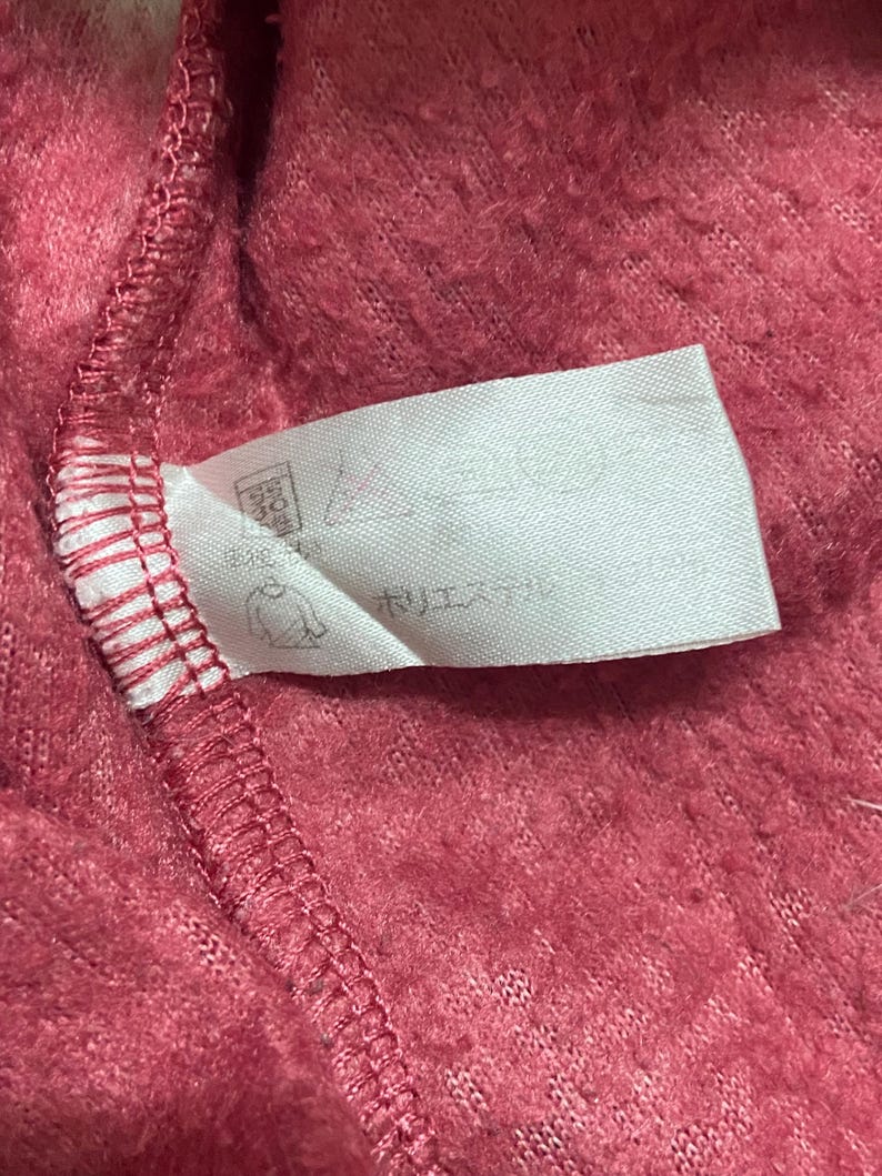 K&ouml;nnte beinhalten: Ein wei&szlig;es Etikett mit schwarzem Text und einer kleinen Grafik einer Person, die sich die H&auml;nde w&auml;scht. Das Etikett ist an einem rosa, flauschigen Stoff befestigt.