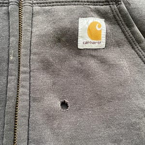 Pode incluir: Close-up de um casaco com capuz cinzento com fecho de correr e um patch com o log&oacute;tipo Carhartt. O casaco com capuz tem um buraco vis&iacute;vel perto da zona do bolso e um fecho de correr de metal. O log&oacute;tipo Carhartt &eacute; um quadrado branco com um C amarelo.