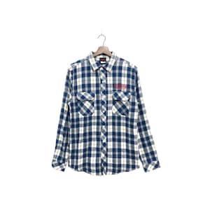 Puede incluir: Una camisa de franela a cuadros azules y blancos con dos bolsillos en el pecho y cuello abotonado. La camisa tiene mangas largas y un logotipo rojo bordado en el pecho izquierdo que dice "Stranger Things". La camisa está colgada en una percha de madera.