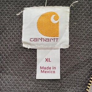 Pode incluir: Close-up de uma etiqueta Carhartt, com um logotipo dourado sobre fundo branco. A etiqueta exibe o nome da marca e o texto "XL Made in Mexico". O fundo &eacute; um tecido cinza texturizado.