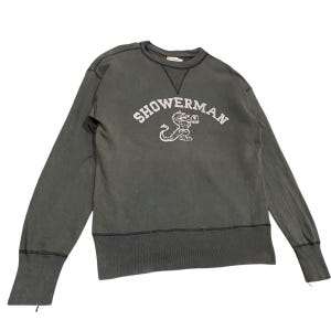 May include: A dark gray sweatshirt with the word "SHOWERMAN" arched across the chest in white lettering. Below the text is a white illustration of a mythical creature. The sweatshirt has long sleeves and a crew neck.