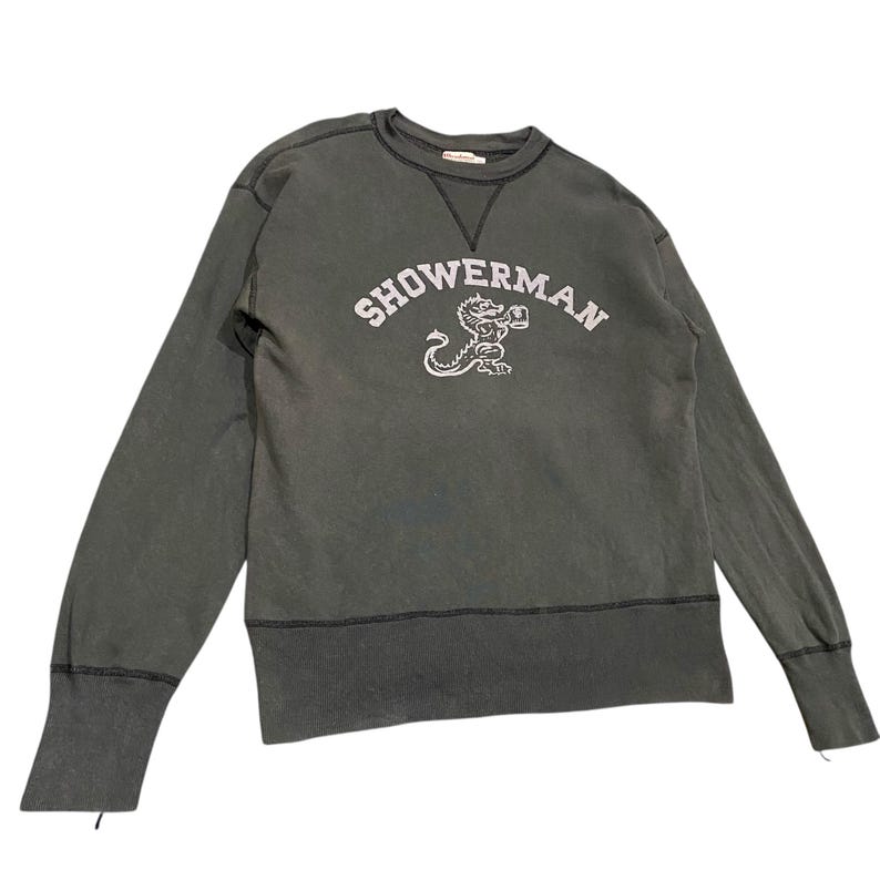 May include: A dark gray sweatshirt with the word "SHOWERMAN" arched across the chest in white lettering. Below the text is a white illustration of a mythical creature. The sweatshirt has long sleeves and a crew neck.