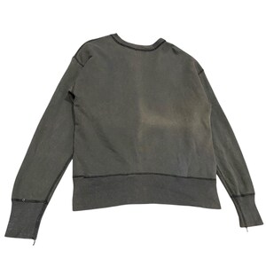 May include: A faded olive green sweatshirt with a crew neck and long sleeves. The cuffs have a small slit and a button detail. The stitching is visible in black thread along the neckline, cuffs, and hem. The fabric appears worn.