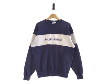 May include: A navy blue sweatshirt with a white stripe across the chest. The stripe has the text "Sergio Tacchini" embroidered in purple.