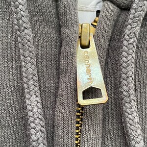 Pode incluir: Close-up de um moletom cinza com z&iacute;per Carhartt. O z&iacute;per &eacute; dourado com o logotipo Carhartt. O moletom tem um cord&atilde;o tran&ccedil;ado ao longo do z&iacute;per e ao redor do capuz.