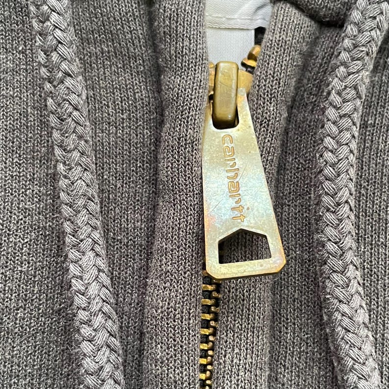 Pode incluir: Close-up de um moletom cinza com z&iacute;per Carhartt. O z&iacute;per &eacute; dourado com o logotipo Carhartt. O moletom tem um cord&atilde;o tran&ccedil;ado ao longo do z&iacute;per e ao redor do capuz.