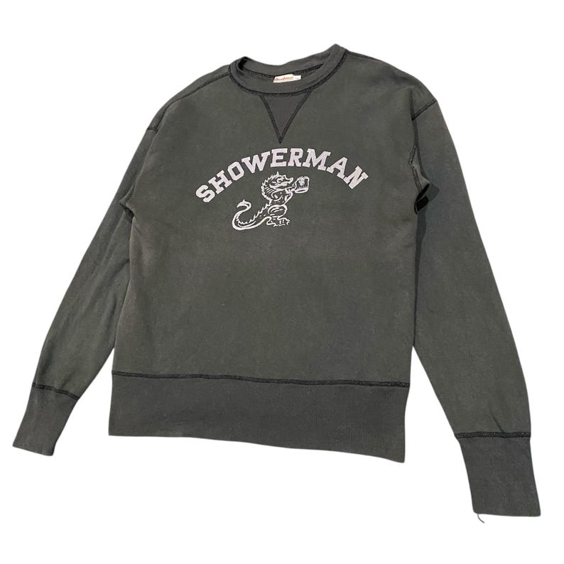 May include: Dark gray sweatshirt with the word "SHOWERMAN" in white arched lettering above a white cartoon dragon. The long-sleeved shirt has a crew neck and ribbed cuffs and hem.