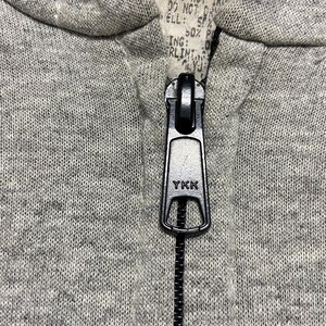 May include: A close-up of a zipper on a gray hoodie. The zipper is black and has the word "YKK" printed on it.
