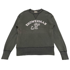 May include: Dark green sweatshirt with the word "SHOWERMAN" in white arched lettering above a white illustration of a dragon-like creature. The sweatshirt has a crew neck, long sleeves, and ribbed cuffs and hem.