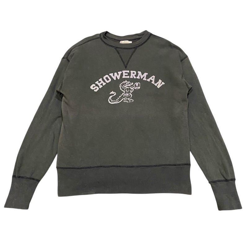 May include: Dark green sweatshirt with the word "SHOWERMAN" in white arched lettering above a white illustration of a dragon-like creature. The sweatshirt has a crew neck, long sleeves, and ribbed cuffs and hem.