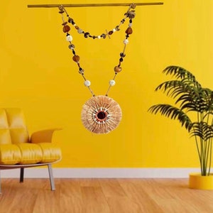 Peut inclure: Une guirlande de perles brunes et blanches avec un élément décoratif rond et tissé qui pend. La guirlande est suspendue à une baguette en bois contre un mur jaune.