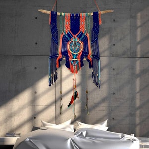 Peut inclure: Une suspension murale en macramé bleu, orange et vert avec une baguette en bois en haut. La suspension présente un motif circulaire au centre et des glands qui pendent du bas.