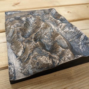 May include: A rectangular, topographical relief map with a detailed, textured surface. The map features shades of brown, gray, and blue, suggesting mountainous terrain. The object is set against a wooden background.
