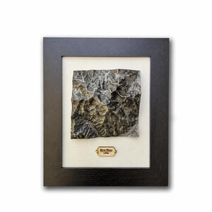 Könnte beinhalten: Gerahmte Reliefkarte des Mont Blanc mit detaillierter, strukturierter Oberfläche in Grau- und Brauntönen. Das Kunstwerk befindet sich vor einem cremefarbenen Hintergrund in einem dunklen Holzrahmen. Ein kleines Schild trägt die Aufschrift "Mont Blanc 4.809m."