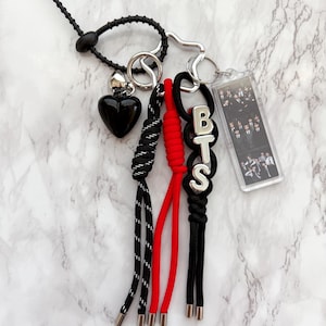 Pode incluir: Um porta-chaves com um pendente de coração preto, um pendente retangular de acrílico com uma fotografia de grupo e três fios de paracord atados em preto, vermelho e preto com pintas brancas. O fio preto inclui contas de letras prateadas BTS.