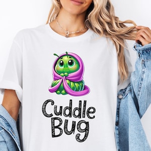 Puede incluir: Camiseta blanca con una oruga verde de dibujos animados que lleva una manta con capucha rosa. El texto "Cuddle Bug" está impreso en negro debajo de la oruga.