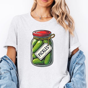 K&ouml;nnte beinhalten: Ein wei&szlig;es T-Shirt mit einem Aufdruck eines Glasbeh&auml;lters gef&uuml;llt mit gr&uuml;nen Gurken und einem roten Deckel. Der Beh&auml;lter hat ein wei&szlig;es Etikett mit der Aufschrift "PICKLES".