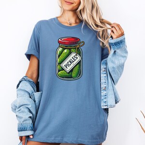 K&ouml;nnte beinhalten: Ein blaues T-Shirt mit einem Aufdruck eines Glases mit Gurken. Das Glas hat einen roten Deckel und ein Etikett mit der Aufschrift "PICKLES".