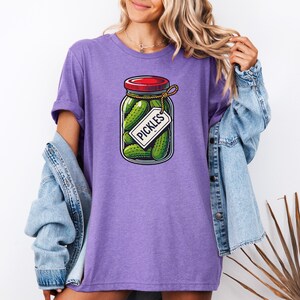 K&ouml;nnte beinhalten: Lilafarbenes T-Shirt mit einem Aufdruck eines Glases mit Gurken, einem roten Deckel und einem Etikett mit der Aufschrift "Pickles".