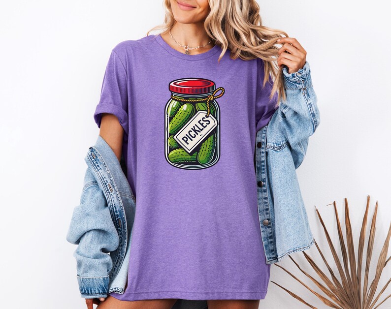 K&ouml;nnte beinhalten: Lilafarbenes T-Shirt mit einem Aufdruck eines Glases mit Gurken, einem roten Deckel und einem Etikett mit der Aufschrift "Pickles".