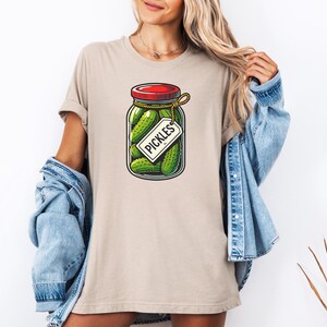 K&ouml;nnte beinhalten: Ein beiges T-Shirt mit einem Aufdruck eines Glasbeh&auml;lters gef&uuml;llt mit gr&uuml;nen Gurken. Der Beh&auml;lter hat einen roten Deckel und ein wei&szlig;es Etikett mit der Aufschrift "Pickles".