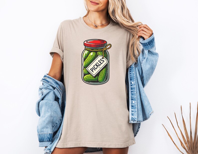 K&ouml;nnte beinhalten: Ein beiges T-Shirt mit einem Aufdruck eines Glasbeh&auml;lters gef&uuml;llt mit gr&uuml;nen Gurken. Der Beh&auml;lter hat einen roten Deckel und ein wei&szlig;es Etikett mit der Aufschrift "Pickles".