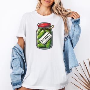 K&ouml;nnte beinhalten: Wei&szlig;es T-Shirt mit einem Aufdruck eines Glasbeh&auml;lters gef&uuml;llt mit gr&uuml;nen Gurken und einem roten Deckel. Der Beh&auml;lter hat ein wei&szlig;es Etikett mit der Aufschrift "PICKLES".