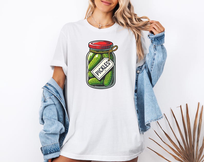 K&ouml;nnte beinhalten: Wei&szlig;es T-Shirt mit einem Aufdruck eines Glasbeh&auml;lters gef&uuml;llt mit gr&uuml;nen Gurken und einem roten Deckel. Der Beh&auml;lter hat ein wei&szlig;es Etikett mit der Aufschrift "PICKLES".
