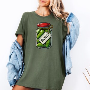 K&ouml;nnte beinhalten: Ein gr&uuml;nes T-Shirt mit einer Cartoon-Illustration eines Einmachglases mit Gurken, einem roten Deckel und einem Etikett mit der Aufschrift "Pickles".