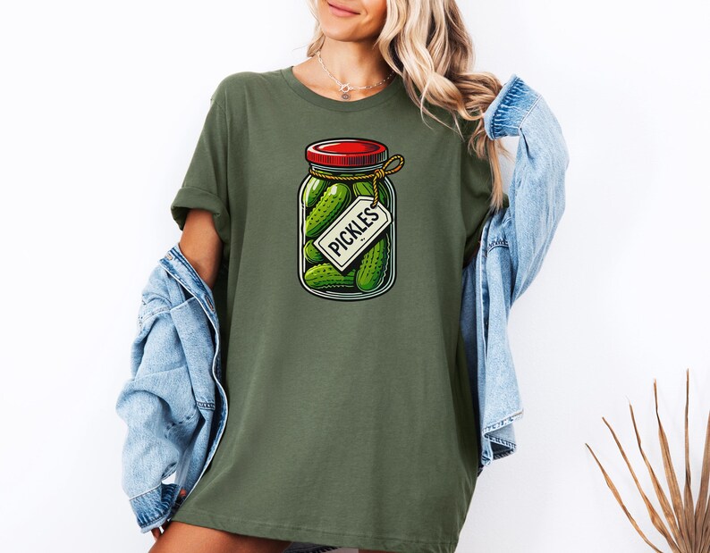 K&ouml;nnte beinhalten: Ein gr&uuml;nes T-Shirt mit einer Cartoon-Illustration eines Einmachglases mit Gurken, einem roten Deckel und einem Etikett mit der Aufschrift "Pickles".