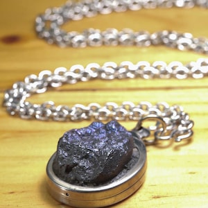Könnte beinhalten: Eine silberfarbene Metall-Halskette mit einem rohen, dunklen, irisierenden Mineralstein in einer ovalen Fassung. Der Anhänger ist an einer silberfarbenen Kette mit ineinandergreifenden runden Gliedern befestigt, die vom Mittelstück wegführen.