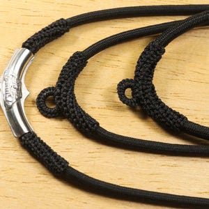 Peut inclure: Gros plan sur un collier en cordon noir avec un fermoir argenté. Le fermoir présente un motif décoratif. Le cordon est noué à plusieurs endroits, créant des boucles. Le collier est présenté sur une surface en bois.