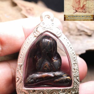 Pode incluir: Uma figura sentada em castanho escuro com as mãos a cobrir o rosto, inserida numa moldura decorativa de pendente em metal prateado. A figura está numa pose meditativa. Uma imagem mostra uma pessoa com vestes e texto em tailandês.