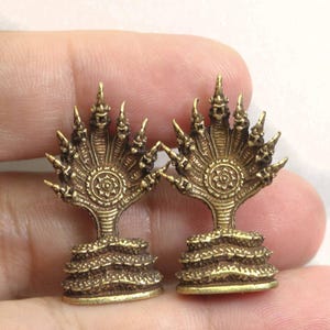 Puede incluir: Dos figuritas de metal de color dorado que representan un naga de siete cabezas, una serpiente mítica de la mitología del sudeste asiático. Cada figurita tiene una base circular con un diseño en relieve.