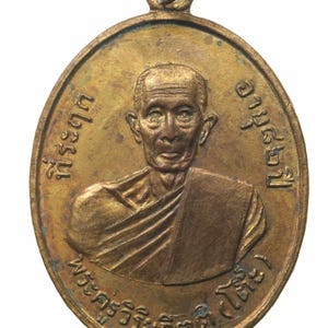 May include: A bronze oval-shaped pendant with a portrait of a man in a monk's robe. The pendant has Thai text around the portrait that reads "Phra Sri Ariyachaikun" and "Luang Pu Suan"