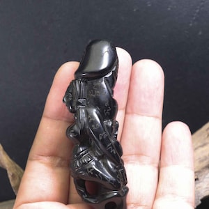 Può includere: Una statuetta nera scolpita di una persona con un cappello. La statuetta è tenuta in mano e presenta dettagli intricati. La figura sembra indossare un cappello e abiti con elementi decorativi. La statuetta ha un foro sul fondo.