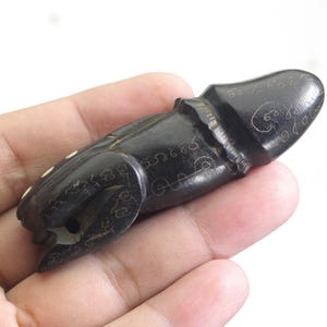 Può includere: Un amuleto di legno intagliato marrone scuro con dettagli incisi e accenti bianchi. L'amuleto ha una forma fallica ed è tenuto nella mano di una persona. La superficie dell'amuleto è liscia e lucida, con intagli intricati.