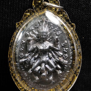 Può includere: Ciondolo ovale raffigurante una divinità argentata con molte braccia e un'acconciatura fluente. Il ciondolo è racchiuso in plastica trasparente con un bordo in filigrana dorata e un anello d'argento per appenderlo. Oggetto religioso.