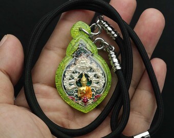 Puede incluir: Un colgante de Buda verde y dorado con un collar de cordón negro. El colgante presenta una imagen detallada de un Buda sentado en posición de loto con un halo alrededor de su cabeza.