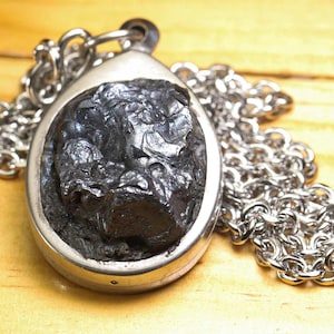 Könnte beinhalten: Ein silberfarbener Metallanhänger mit einem rohen, dunkelgrauen, strukturierten Stein in einer ovalen Fassung. Der Anhänger ist an einer passenden silberfarbenen Metallkette mit abgerundeten Gliedern befestigt, was ein stimmiges Schmuckstück ergibt.