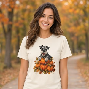 Puede incluir: Una camiseta de manga corta de color crema con un estampado gráfico de un perro Schnauzer negro y gris descansando sobre una pila de calabazas naranjas rodeadas de hojas de otoño. El diseño está centrado en el pecho de la prenda.