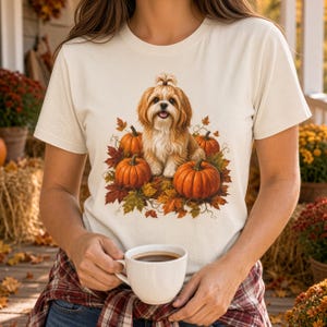 May include: A cream-colored t-shirt featuring a graphic print of a long-haired brown and white dog sitting among orange pumpkins and colorful autumn leaves. The dog has a small topknot hairstyle and is positioned centrally on the chest area.