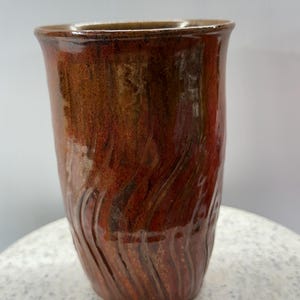 May include: A handmade ceramic tumbler with a glossy, reddish-brown glaze. The tall, cylindrical cup features a slightly flared rim and vertical, wave-like textured patterns on the lower half. The cup is approximately 15 cm tall.