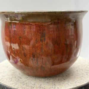 May include: A ceramic bowl with a rounded shape and a wide opening. The bowl has a rich, reddish-brown glaze with darker, mottled accents. The rim has a contrasting, lighter brown colour. This decorative bowl is suitable for various uses.