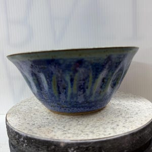 May include: A handmade ceramic bowl with a blue glaze and textured vertical lines. The bowl has a wide, flared shape and a light brown rim. The bowl is sitting on a gray and black surface.