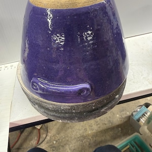 May include: A handmade ceramic pot with a deep purple glaze. The pot has a rounded shape with a textured surface and a light tan rim. A handle is attached to the side. The base is a dark gray color.