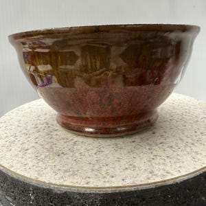 May include: A handcrafted ceramic bowl with a glossy glaze. The bowl displays a rich colour gradient, shifting from a dark brown rim to a reddish-brown base. The bowl is round with a slightly flared rim and a pedestal base.